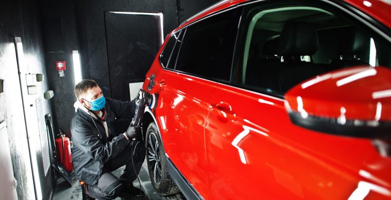 Car detailing concept. Man in face mask with orbital polisher in repair shop polishing orange suv car.